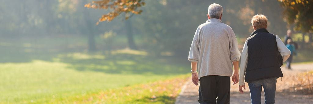A retired couple walks together on a tree-lined park trail.